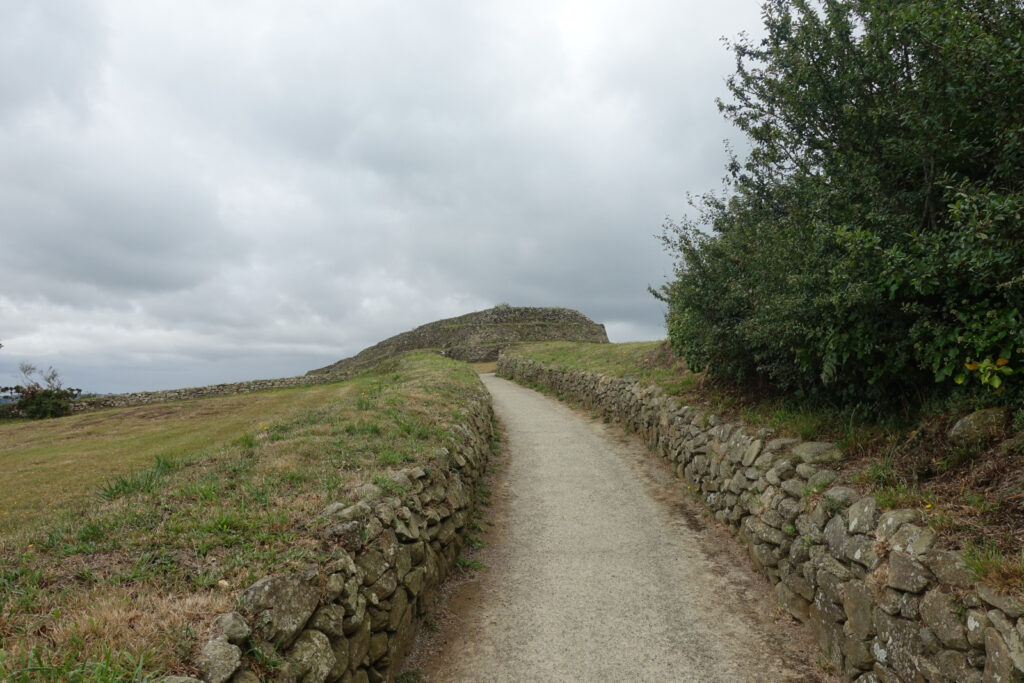 Weg zum Grabhügel Cairn de Barnenez. Der Weg ist umgeben von Trockensteinmauern. Am Ende des Wegs ist der Grabhügel aus Steinen zu erkennen.