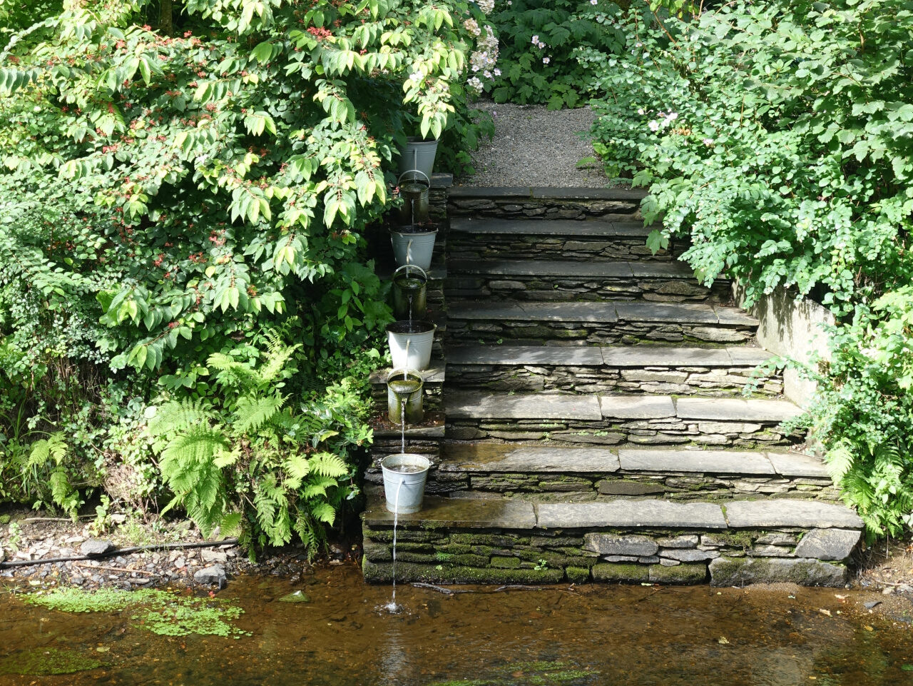 Ein Brunnen auf einer Treppe, auf jeder Stufe eine Giesskanne wo das Wasser jeweils in die nächste rinnt.