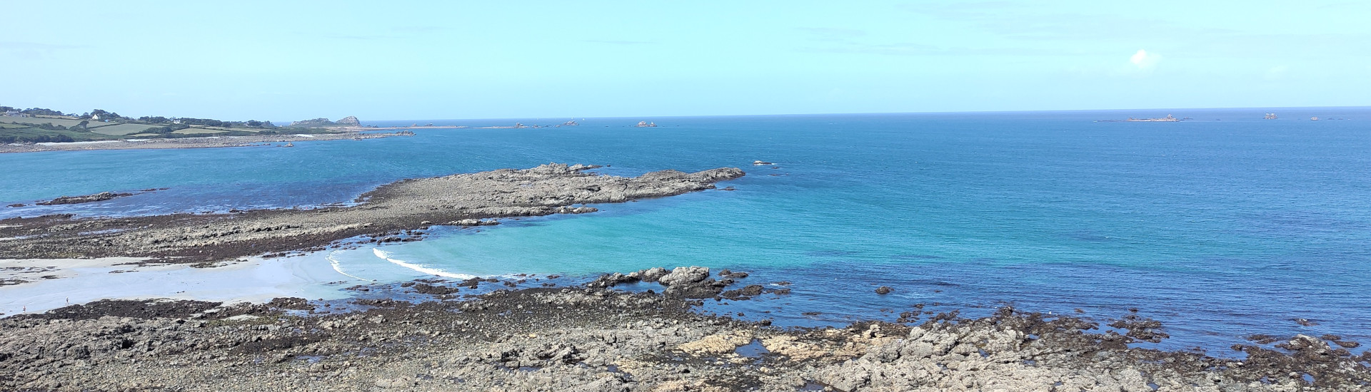 Küstenbild. Felsen umgeben von klarblauem Wasser. Der Himmel ist hellblau.