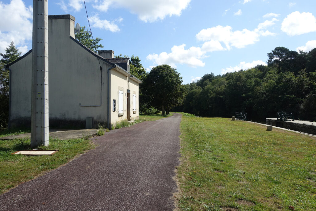 Links ist ein weisses Gebäude aus Beton zu sehen. Das Gebäude dient als Wärterhaus für die Schleuse. Zwischen der Schleuse rechts sind ein schmaler Weg und eine Wiese.