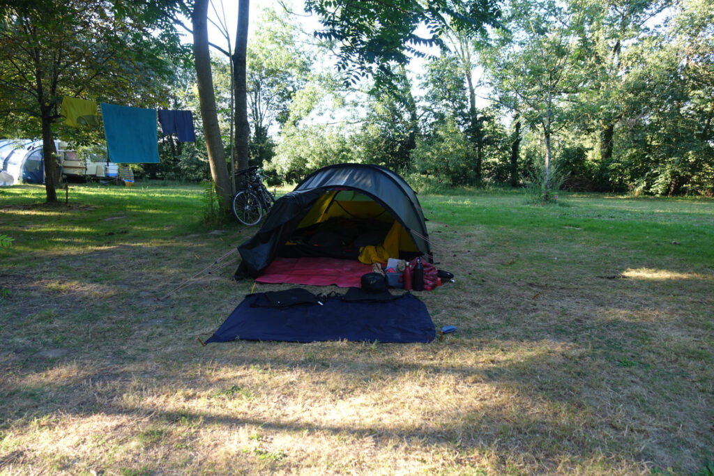 In der Bildmitte ein Zelt mit offener Abside und Picknick-Decke. Ringsherum Bäume und Wiese.