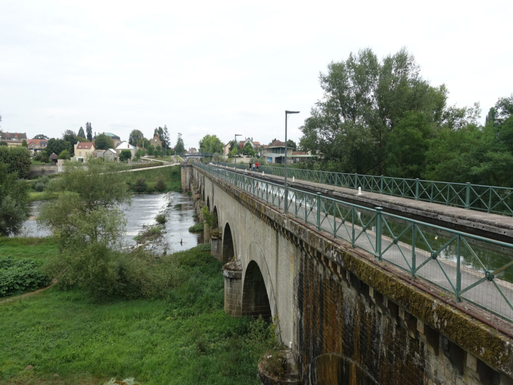 Von der rechten Bildseite verläuft der Kanal hin zur Bildmitte. In der Bildmitte bis zum linken Bildrand ist der Fluss Loire zu sehen, der unter der Brücke hindurch fliesst. Im Hintergrund sind einige Häuserzeilen der Stadt Digeon zu sehen.