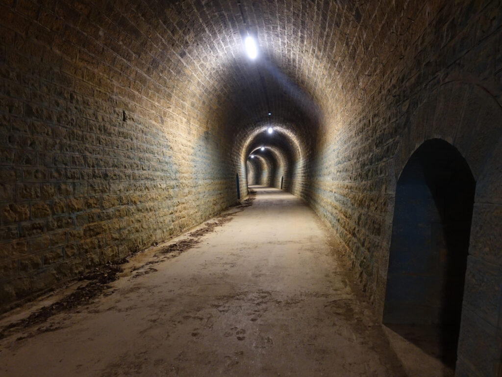 Im innern eines Tunnels. In regelmässigen Abständen gibt es an der Tunneldecke Lampen. Dadurch wird eine Art Lichtbogen im hinteren Tunnelteil erzeugt. Die Tunnelwand ist gemauert mit Natursteinen.