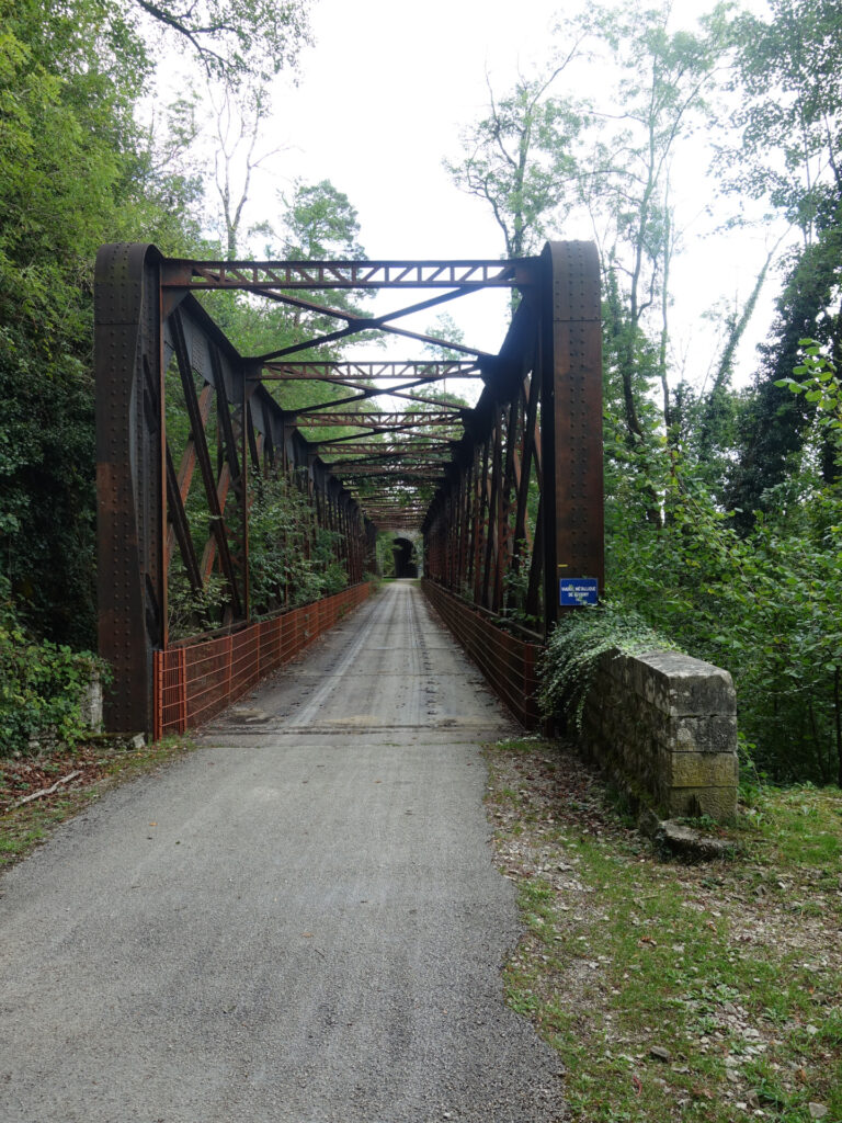 Naturstrasse, die auf eine alte eiserne rostfarbende Eisenbahnbrücke führt. Die Eisenbahnbrücke ist umgeben von Bäumen. Ganz am Ende ist ein Tunneleingang erkennbar.