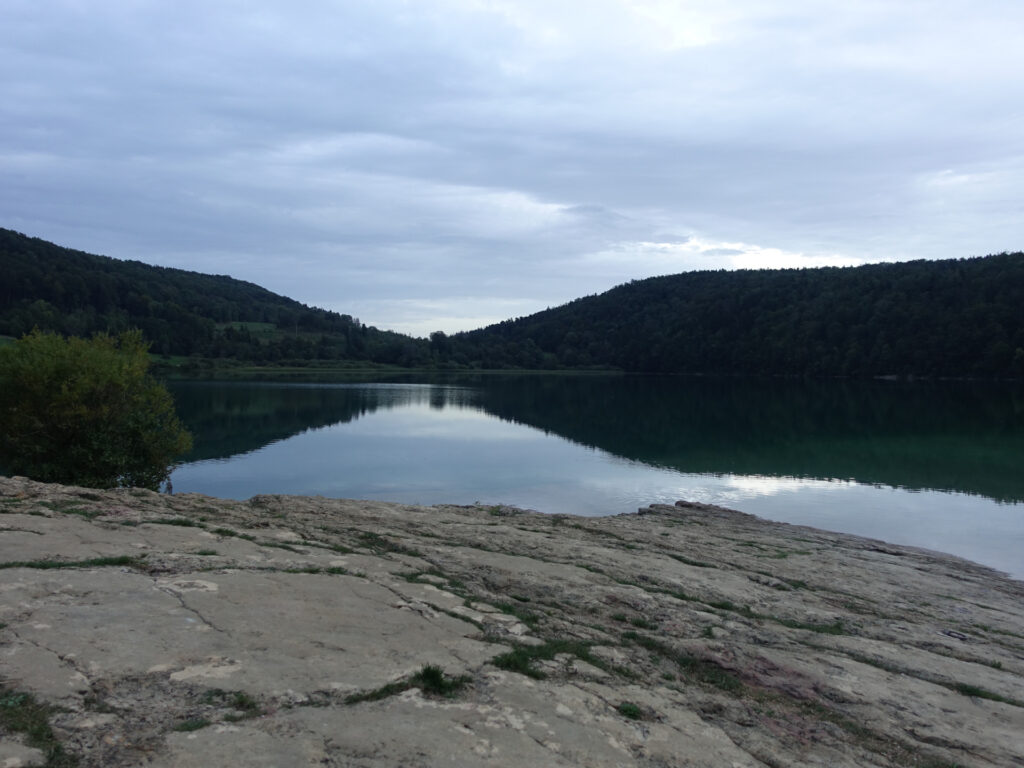 Im Vordergrund ein felsiges Ufer. In der Bildmitte der See, in dessen Wasser sich der bewaldete Hügel im Hintergrund spiegelt.