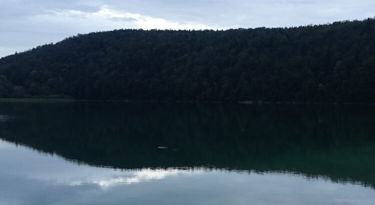 Bild eines Sees. Im Vordergrund Wasser mit Spiegelung der Hügel im Hintergrund.