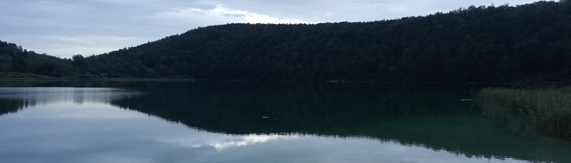 Bild eines Sees. Im Vordergrund Wasser mit Spiegelung der Hügel im Hintergrund.