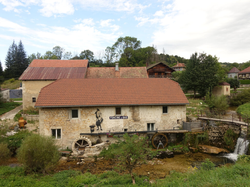 Ein altes Mühlegebäude aus Stein. Die Wasserräder sind noch an den Mauern des Gebäudes angebracht, der Bach fliesst aber nicht mehr über die Räder sondern vor dem Gebäude im Flussbeet. HInter der Mühle sind weitere Häuser des Ortes erkennbar.