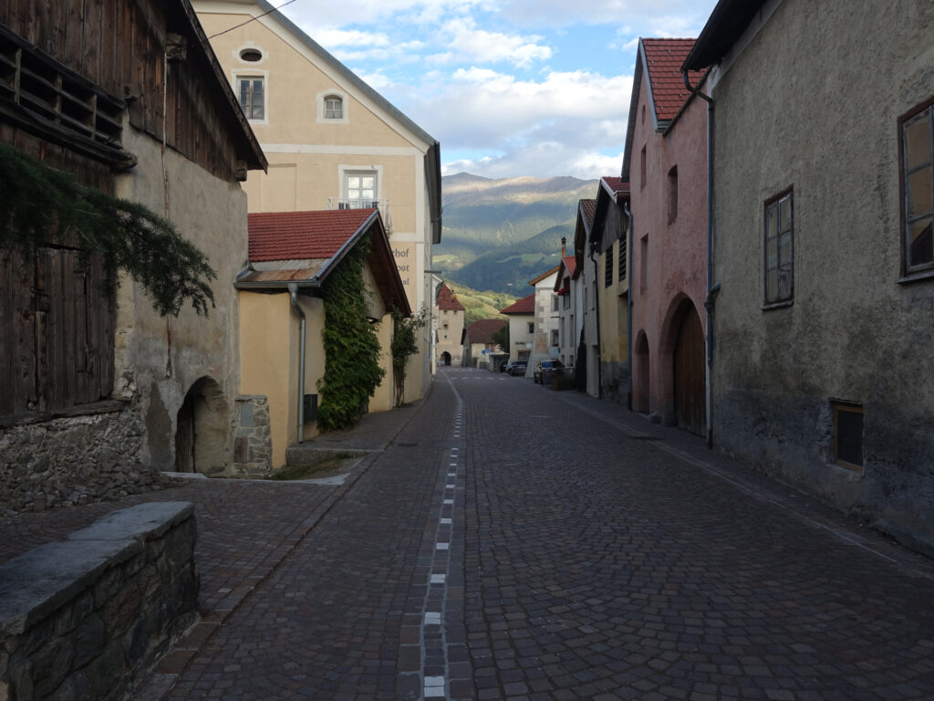 Strasse umgeben von alten Gebäuden. Im Hintergrund sind die Stadtmauer sowie die umgebenden Berge zu erkennen.