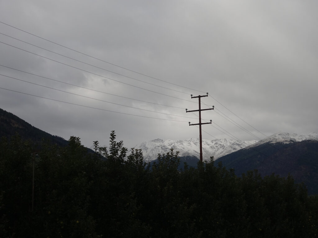 Unter der Wolkendecke sind auf den Berggipfeln im Hintergrund die ersten Schneefelder erkennbar. Im Vordergrund steht ein alter Strommast.
