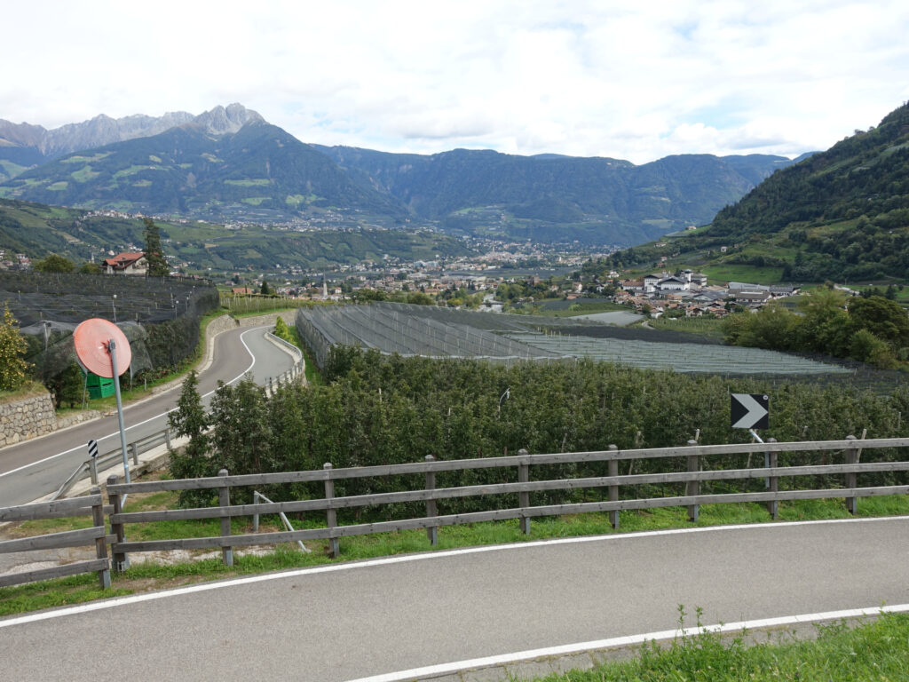 Blick von einem Ausblickpunkt über die Wein- und Apfelplantagen nach Merano. Im Hintergrund sind die weitere Bergketten erkennbar.