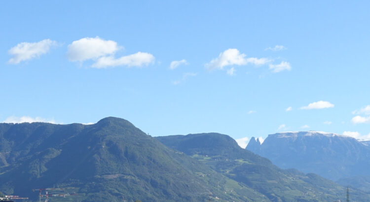 Hinter einem grünen Hügelkamm sind bei strahlendem Sonnenschein unter dem blauen Himmel die Ausläufer der Dolomiten um Bolzano erkennbar.