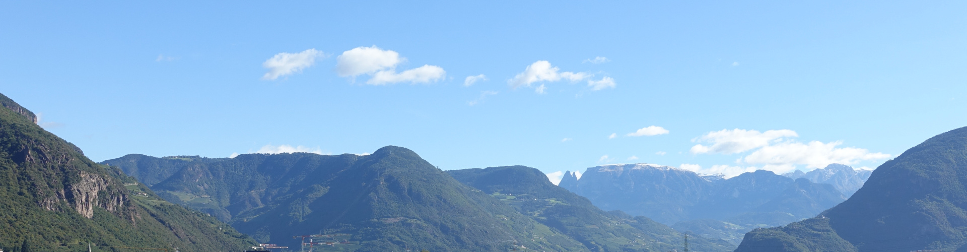 Hinter einem grünen Hügelkamm sind bei strahlendem Sonnenschein unter dem blauen Himmel die Ausläufer der Dolomiten um Bolzano erkennbar.