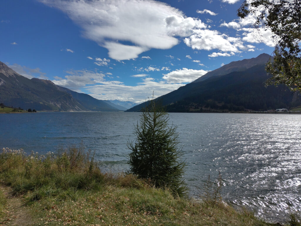 Blick vom Ufer auf den Stausee Reschen. Im Hintergrund Bergflanken, blauer Himmel und weisse Wolken.