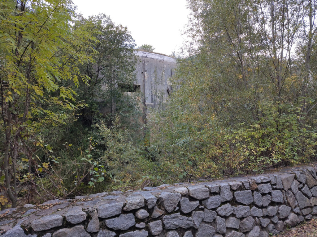 Hinter einer kleinen Bruchsteinmauer, Gebüsch und Bäumen ist ein Bunker aus Beton aus dem 2. Weltkrieg zu erkennen.