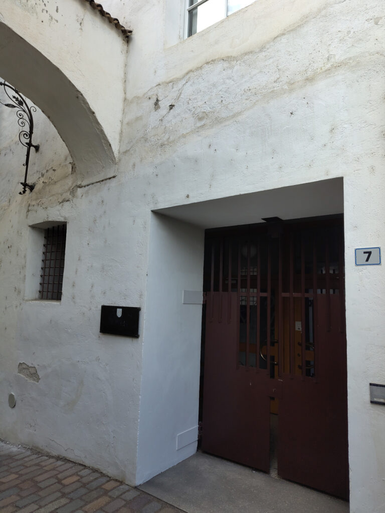 Altes Stadtgebäude mit weissen Mauern. Auf der höhe des ersten Stockwerkes führ ein Bogen zum vis-à-vis liegenden Gebäude. Die Tür zum Stadtarchiv ist aus Massivholz. Links davon ist das Metallschild mit dem Namen des Archivs angebracht. Noch etwas weitere links ist ein kleines vergittertes Fenster.