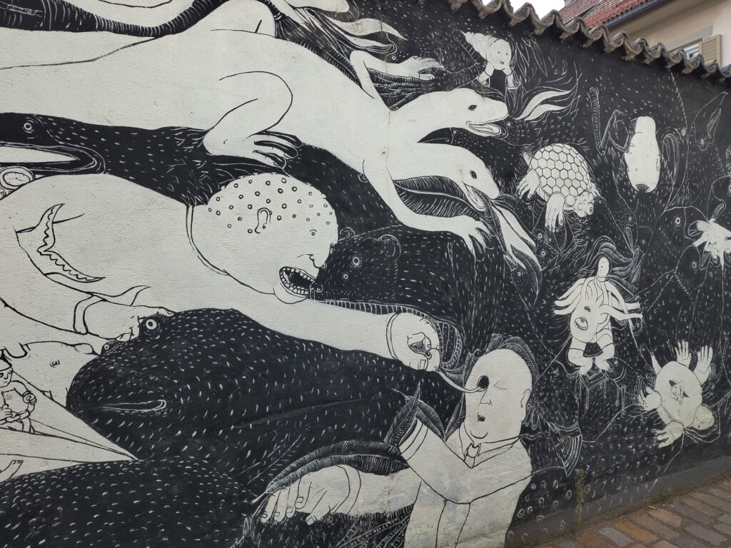 Schwarzweisses Wandgemälde. Abgebildet sind verschiedene Fabelwesen unter anderem eine zweiköpfige weisse Echse. Die anderen hellen Fabelwesen erinnern an Schildkröten, Krebse und Tintenfische. Die dunklen Tiere erinnern an Vögel und Nagetiere. Im unteren Bildteil zieht ein menschlich aussehendes Wesen einem anderen das Auge aus dem Kopf.