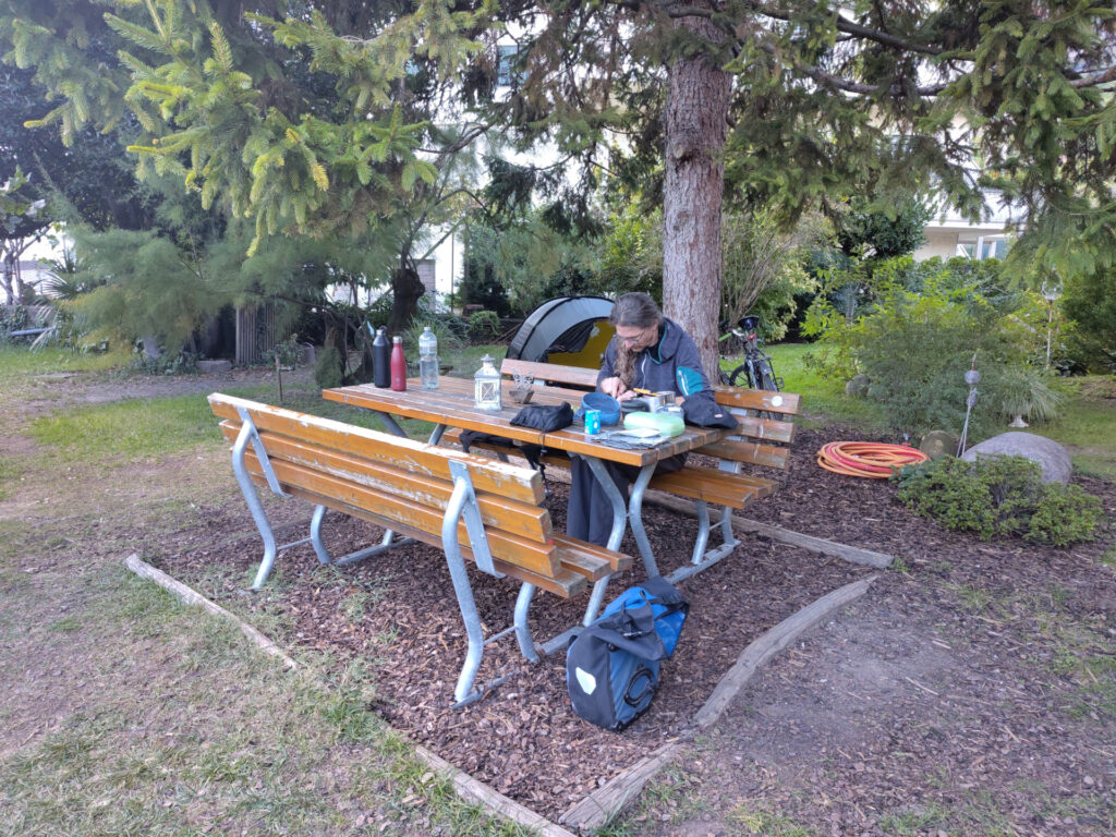 Unser Zeltplatz im Garten vom Camping Obstgarten. Unter einem Baum hat es einen Sitzplatz mit Bank. Eine Person rüstet Gemüse. Rund um die Person hat es Kochutensilen für die Campingküche. Im Hintergrund ist ein grünes Tunnelzelt erkennbar.