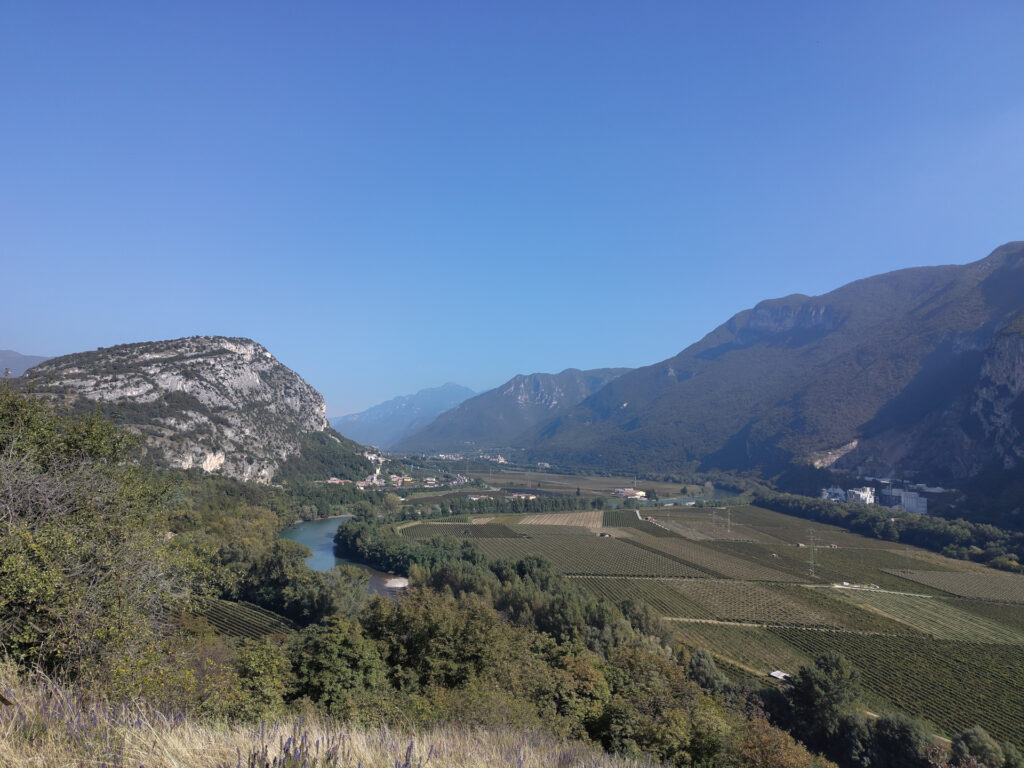 Blick über das Etschtal von einer Anhöhe. In der Ebene sind Ackerland und Weinplantagen. Rechts und links die Bergketten des Südtirols.