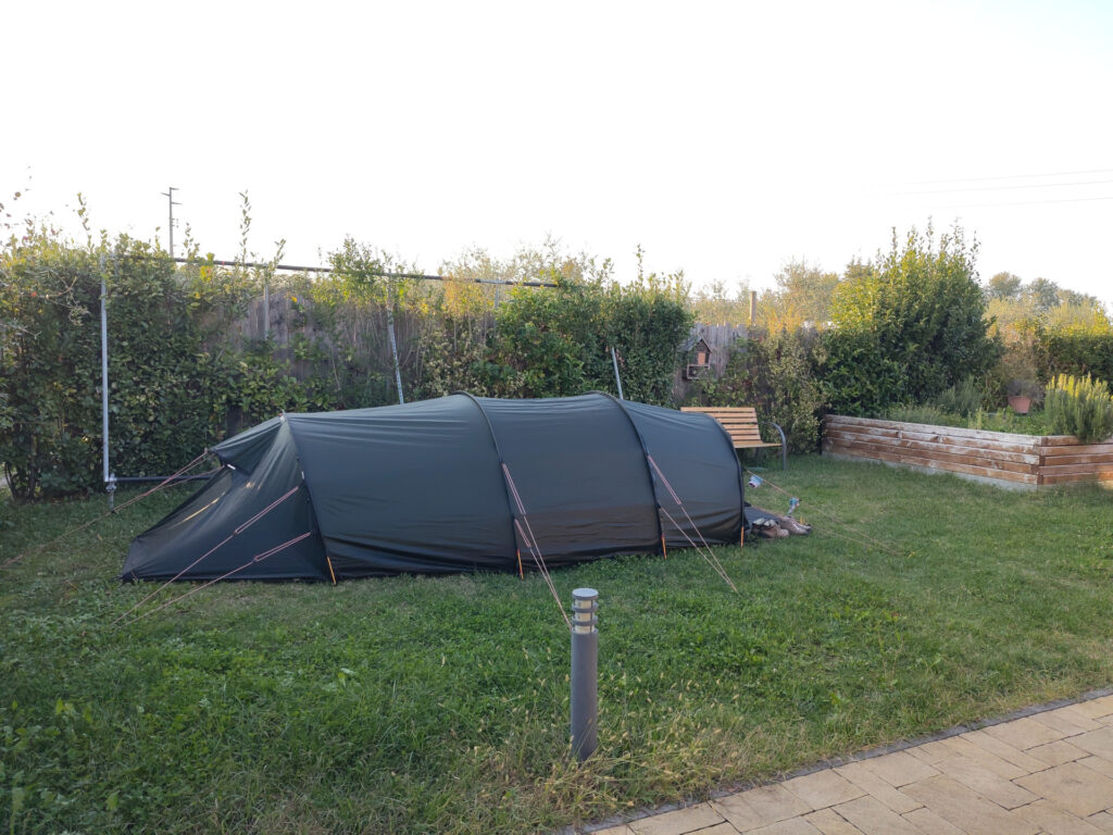 Ein Tunnelzelt auf einem Rasen. Hinter dem Zelt hat es eine Hecke und rechts neben dem Zelt ein Hochbeet mit Kräutern.